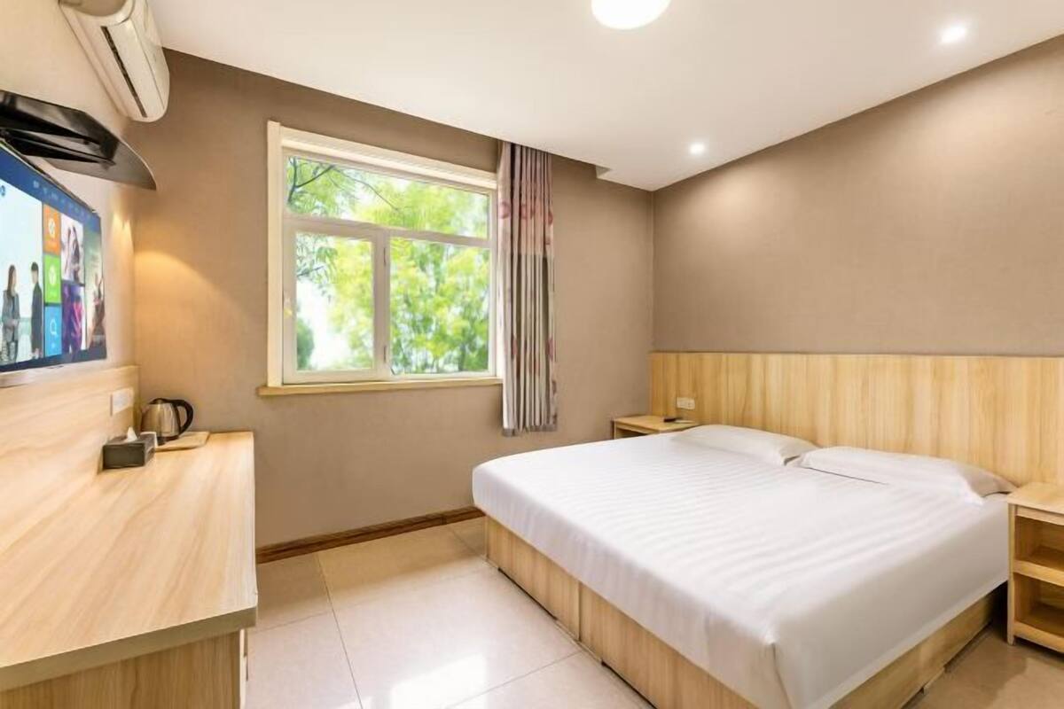 A hotel room with a bed, a wooden nightstand, a desk with a kettle, a window with curtains, and an air conditioning unit.