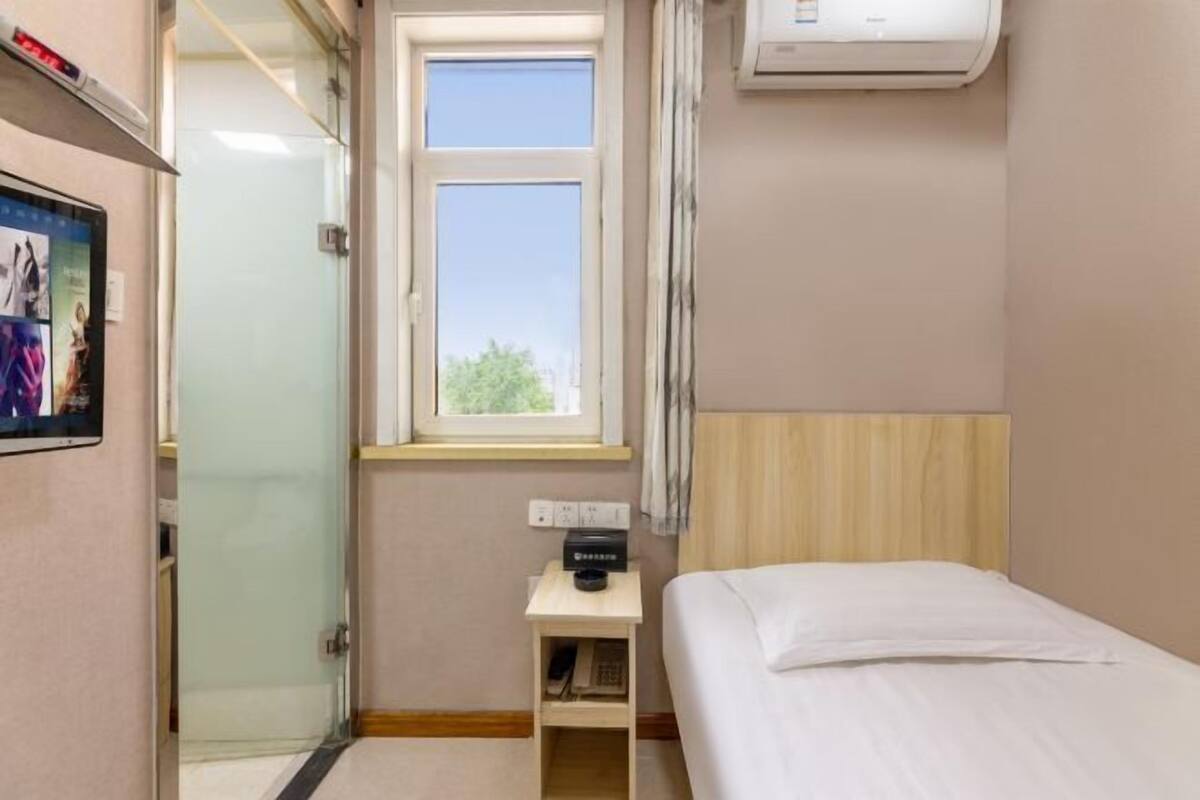 A hotel room with a bed, a bedside table with a lamp, a window, and a wall-mounted air conditioner.