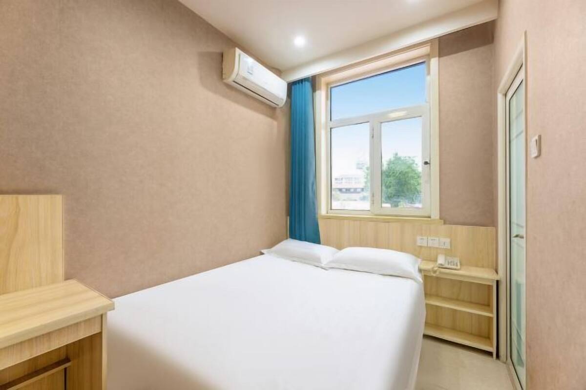 A hotel room with a bed, a desk, a window with curtains, and an air conditioning unit.