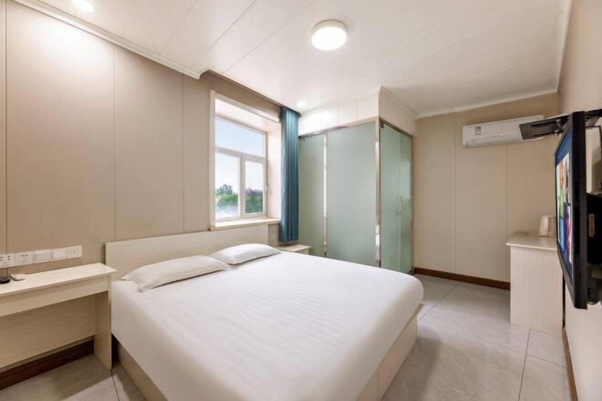 A hotel room with a bed, bedside table, window, wardrobe, and an air conditioning unit.