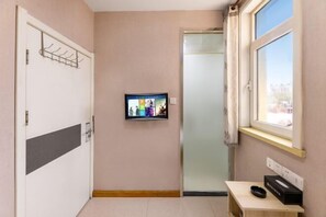 Quarto com porta, um cabide, televisão na parede, janela e uma pequena mesa com uma caixa e um controle remoto.
