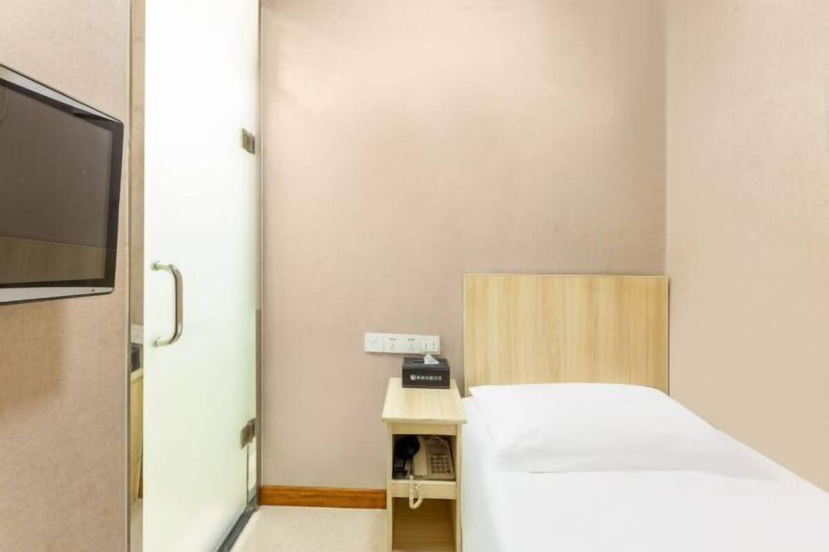 A small, single-bed room with a wooden headboard, a bedside table with a phone and a box, and a wall-mounted TV.