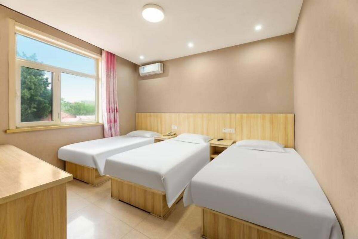 A hotel room with two single beds, a desk, a window with a view, and an air conditioning unit.