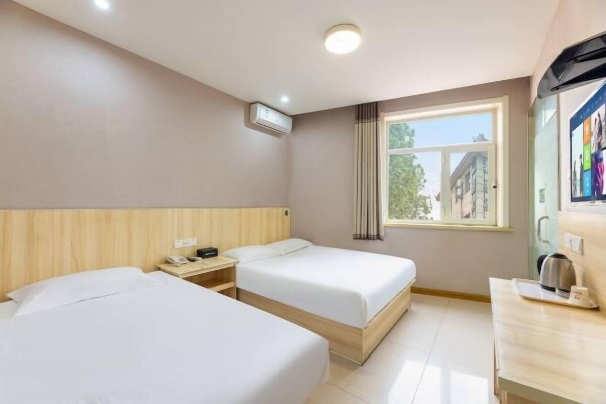 A hotel room with two beds, a small table with a kettle, a window with curtains, and an air conditioning unit on the wall.