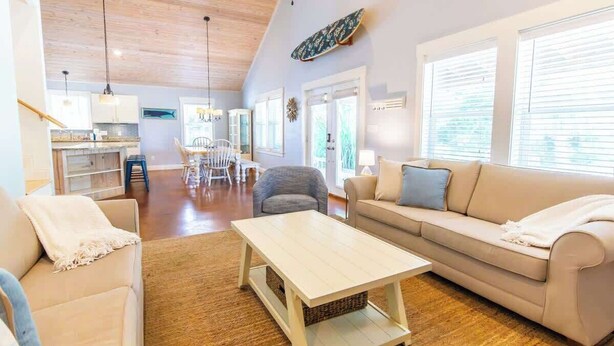 A spacious living area with a sofa, a coffee table, and a dining table. There is a kitchen in the background with a window overlooking the dining area.