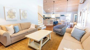 A spacious living area with a beige sofa, a coffee table, and a gray armchair. A kitchen with a central island and bar stools is visible in the background.