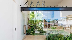 The entrance of KAAB South Beach with a glass door, a logo, and a view of the street and greenery outside.