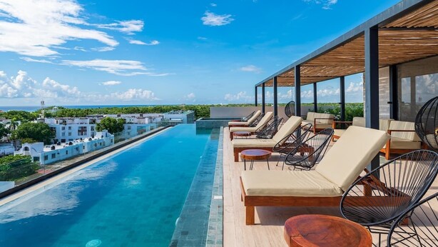 A rooftop infinity pool with lounge chairs and a wooden pergola, overlooking a coastal area.