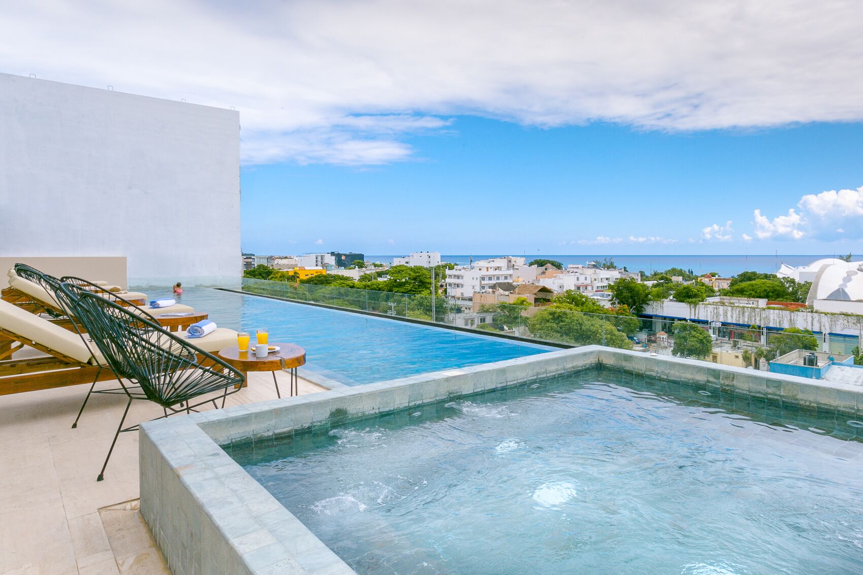 Piscina infinita en la azotea con sillones de descanso y una mesa, con vistas a un paisaje urbano costero.