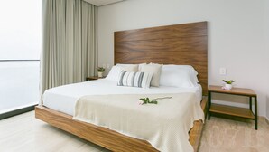A modern bedroom with a wooden headboard, a large bed with white bedding, a bedside table with a plant, and a view of the outdoors.