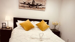 Un lit bien fait, recouvert d’une couvre-lit à motifs géométriques, agrémenté de deux coussins de couleur moutarde et d’un objet décoratif en forme de cœur posé sur le lit.
