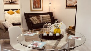 Un salon moderne avec une table à manger en verre entourée de chaises blanches, un canapé foncé agrémenté de coussins et une table basse ornée d’objets décoratifs.