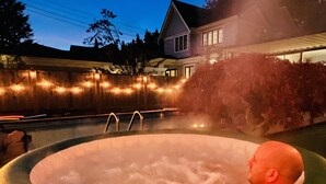 Un bain à remous avec une personne à l’intérieur, entouré d’une clôture en bois et illuminé de guirlandes lumineuses.
