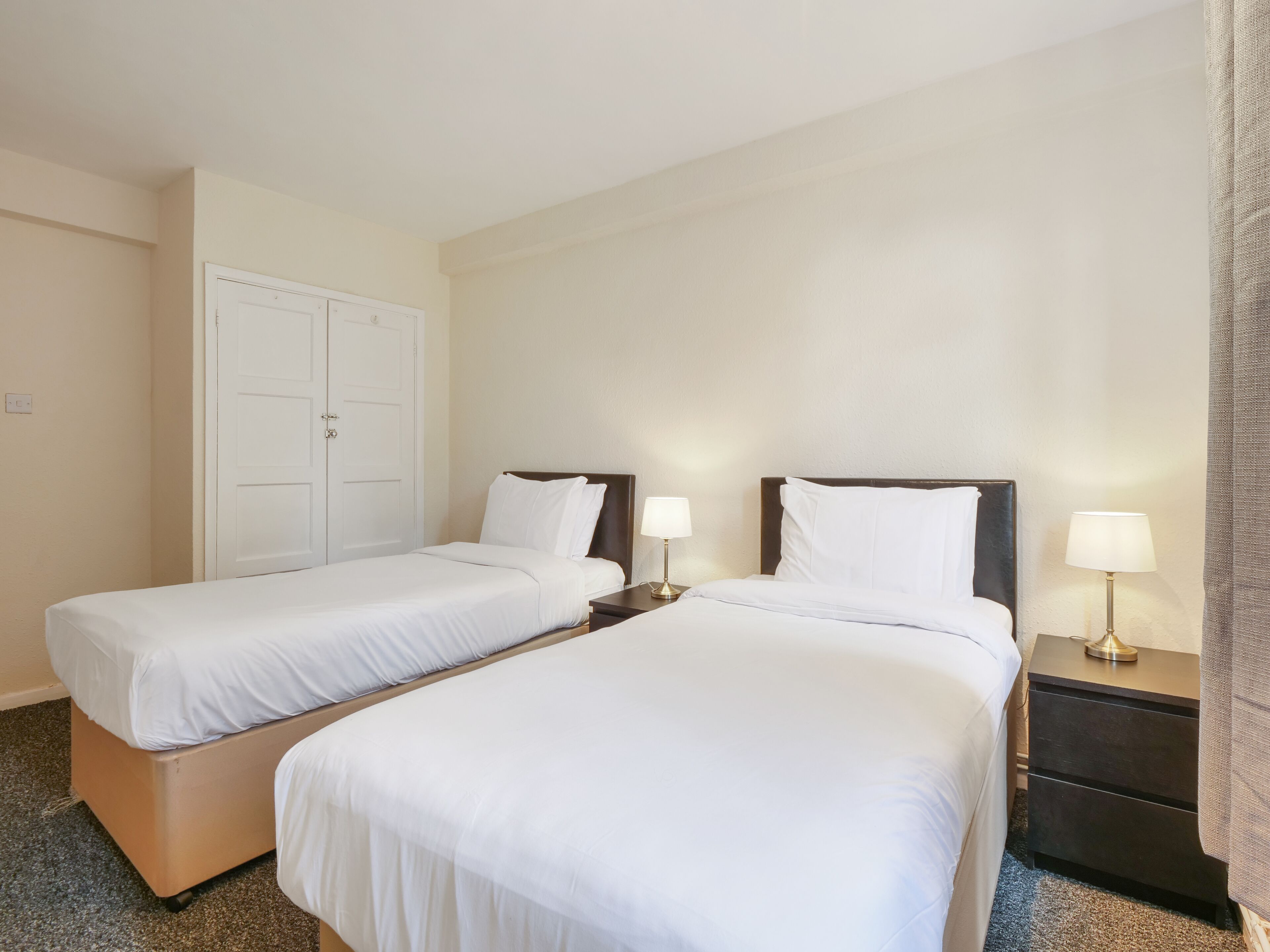 A hotel room with two single beds, a wardrobe, a bedside table with a lamp, and a carpeted floor.