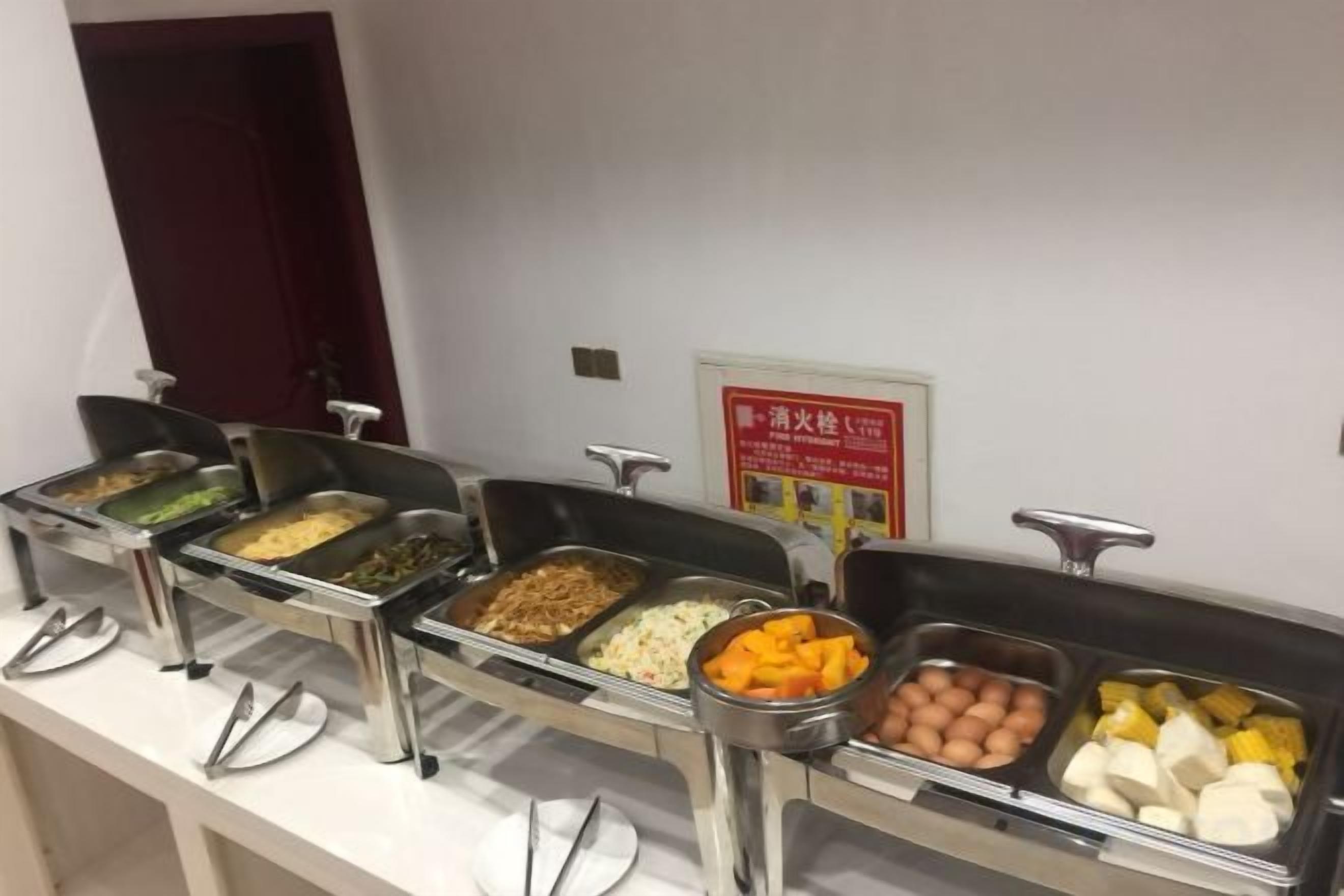 A buffet line with various food items in metal containers, a door in the background, and a sign on the wall.