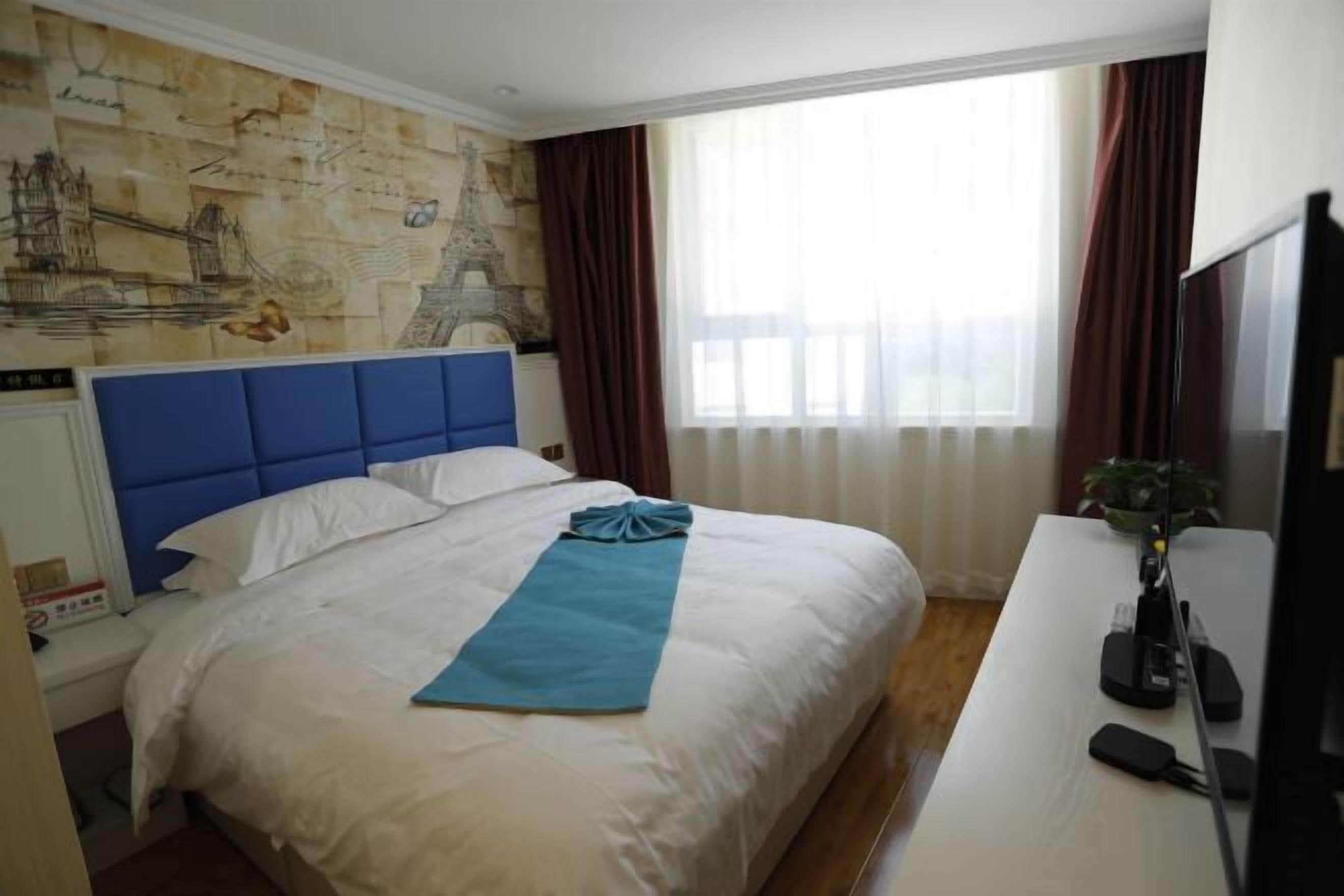 Una camera d'albergo con un letto grande, una testiera blu, una mappa appesa al muro, una scrivania con un computer e una finestra con tende.