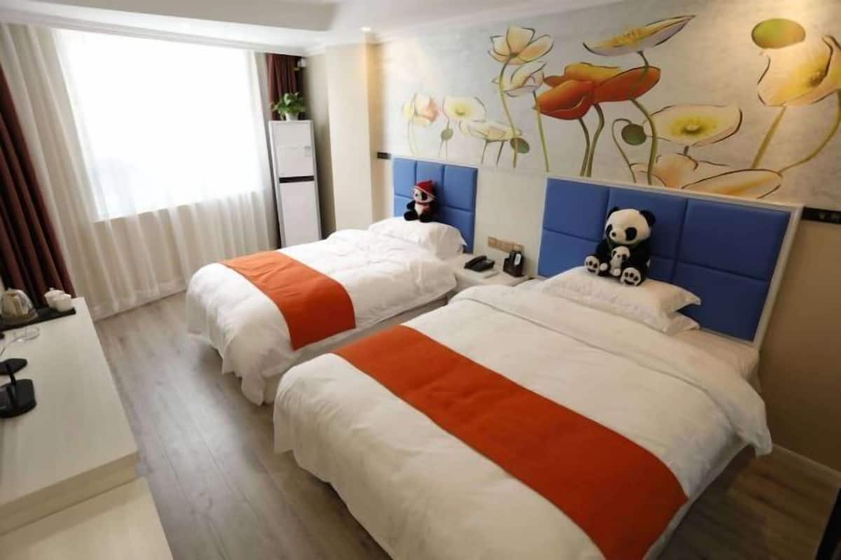 Ein Hotelzimmer mit zwei Betten, einem Blumenbild an der Wand, einem Panda-Plüschtier auf einem Bett und einem kleinen Tisch mit einer Lampe.