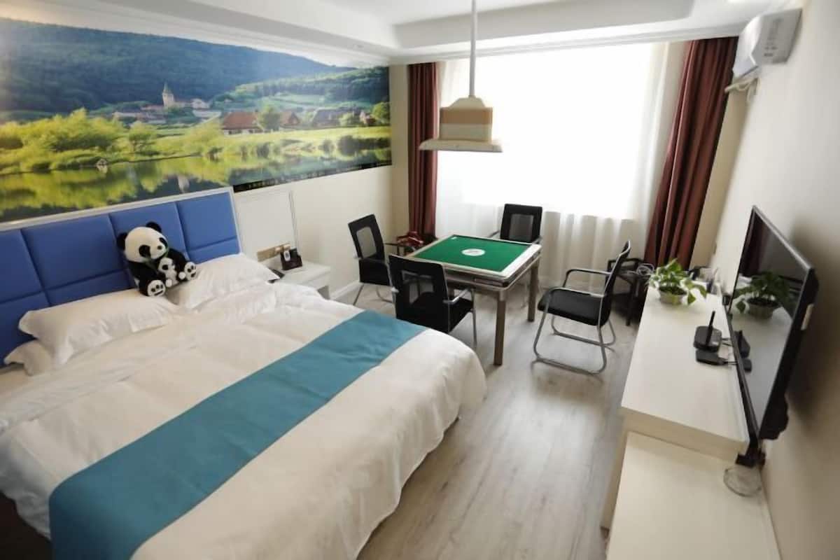 Ein Hotelzimmer mit einem Bett, einem Schreibtisch mit grüner Tischplatte, einem Stuhl, einem Fernseher und einem Fenster mit Vorhängen.