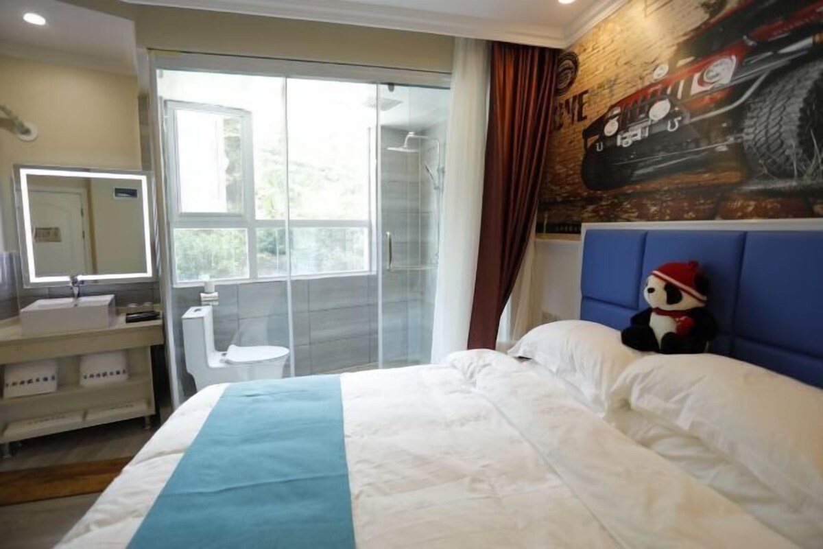 A hotel room with a bed, a bathroom with a shower, a window, and a wall with a mural.