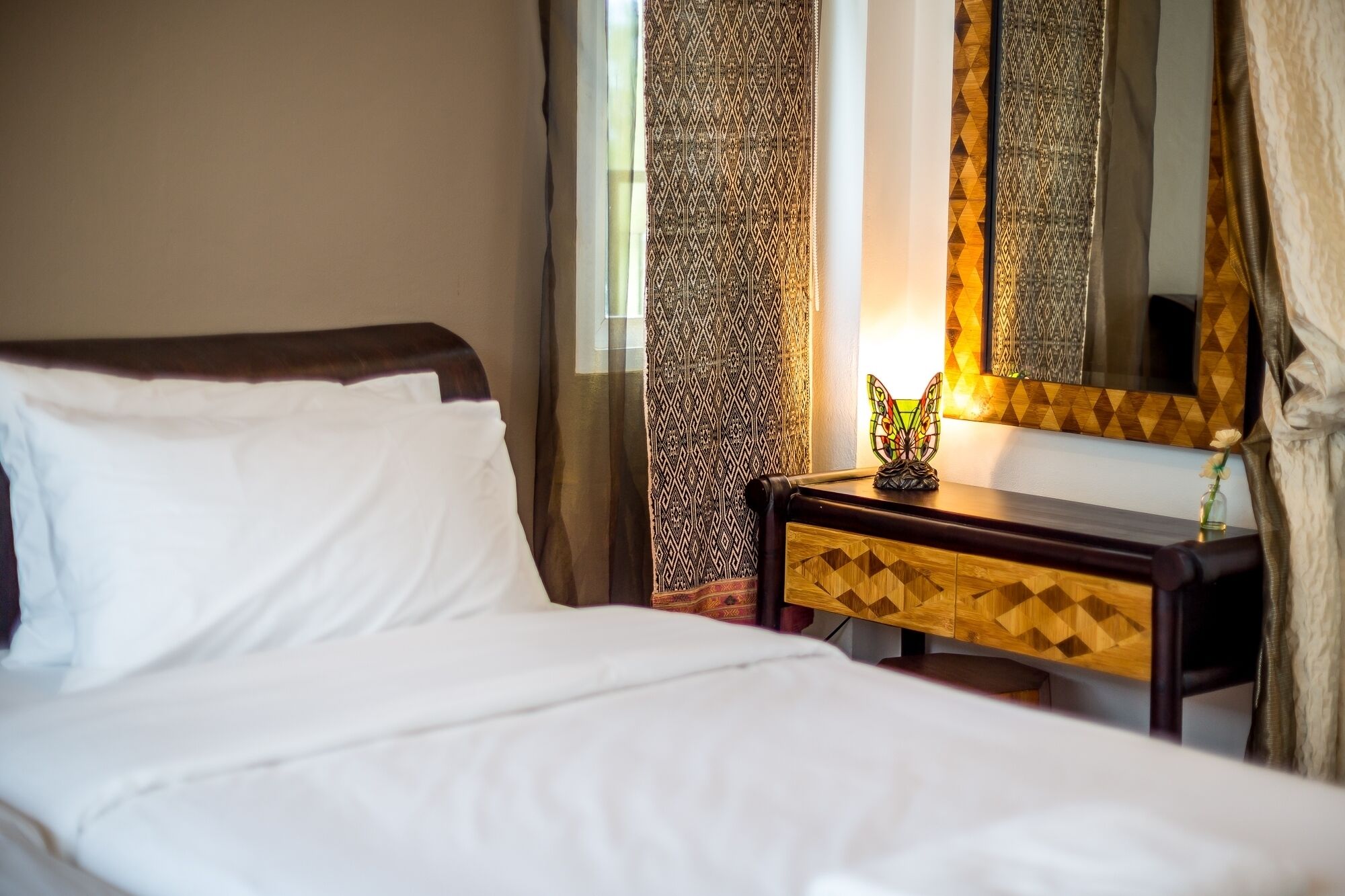 A neatly made bed with white linens, a wooden nightstand with a geometric drawer, a mirror, and a decorative item on the nightstand.