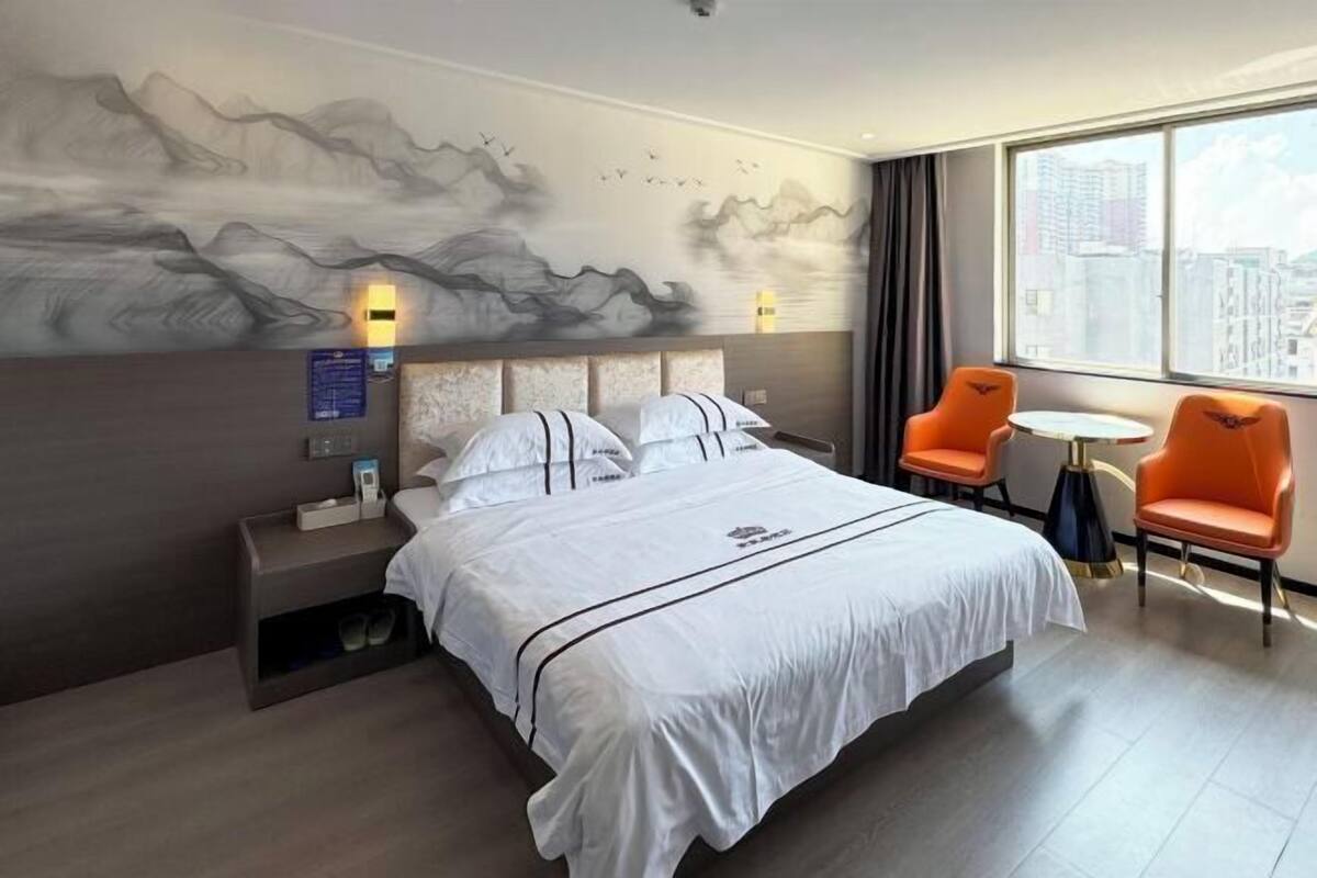 A hotel room with a bed, a nightstand, a small table, two chairs, a wall mural, and a window with a city view.