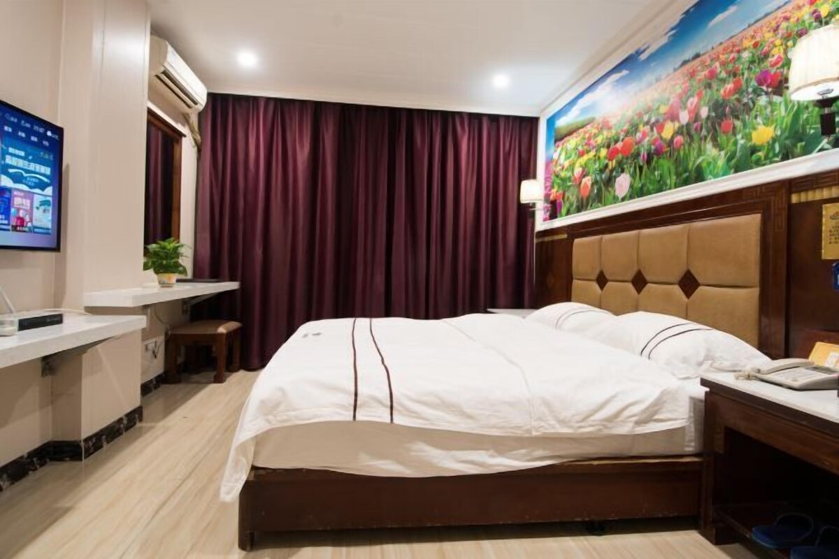 A hotel room with a bed, a desk, a chair, a TV, and a painting of flowers on the wall.