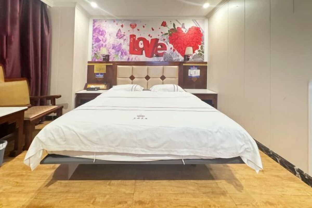 A hotel room with a large bed, a headboard with a decorative design, two bedside tables with lamps, and a wall decoration with the word "Love".