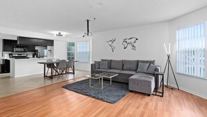Un salon moderne avec un canapé gris, une table basse et un coin repas avec des chaises. On aperçoit au fond une cuisine équipée d’un four à micro-ondes et d’un réfrigérateur.