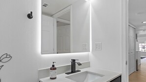 Une salle de bain avec un lavabo blanc, un robinet noir et un grand miroir. Un flacon de savon liquide est posé sur le comptoir.