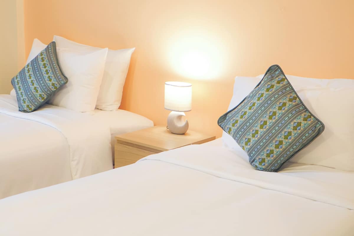 Habitación de hotel con dos camas, ropa de cama blanca, una mesita de noche con lámpara y almohadas estampadas.