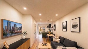 Uma sala de estar moderna com sofá, mesa de centro, área de jantar com cadeiras e um quadro de uma paisagem urbana.