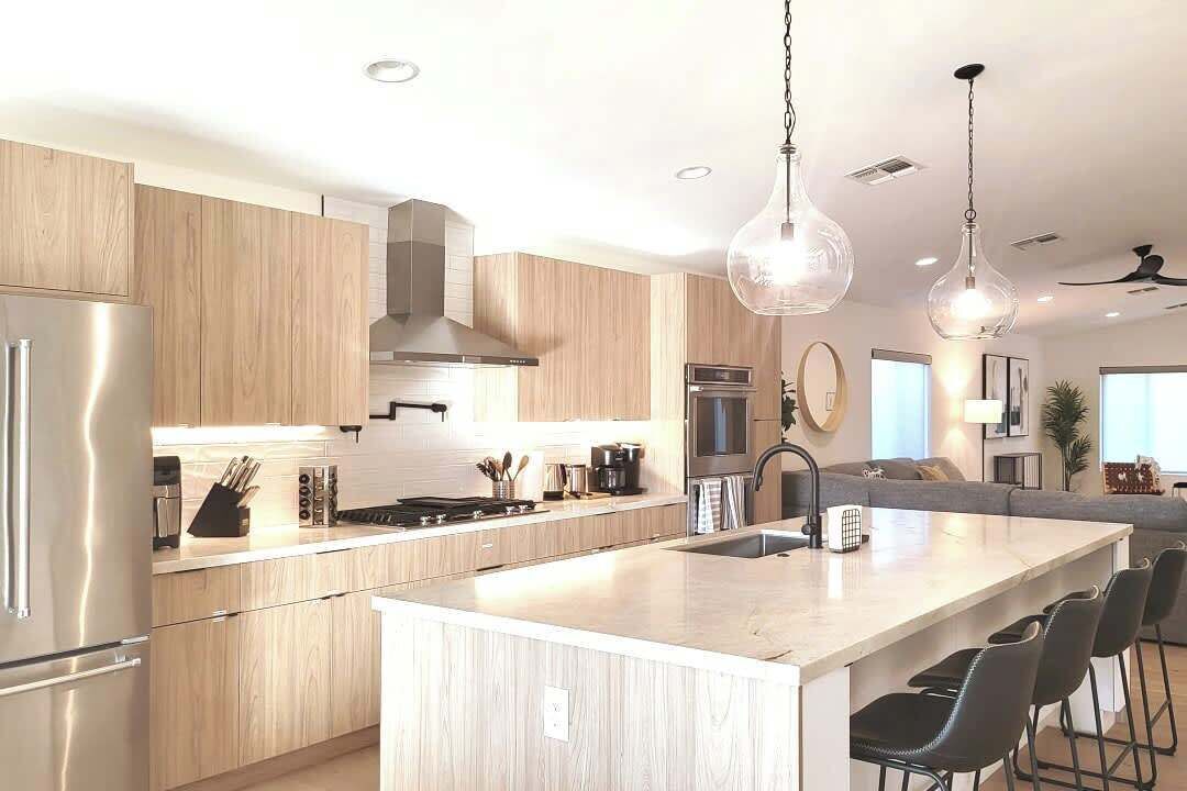 A modern kitchen with wooden cabinets, stainless steel appliances, a central island with bar stools, pendant lighting, and a view into the living area.