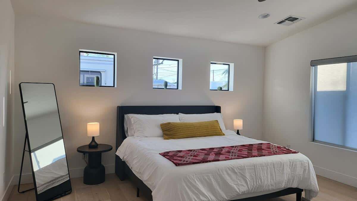 Un dormitorio con cama, mesitas de noche, un espejo y tres ventanas.