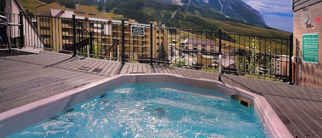 A hot tub with a mountain view, surrounded by a wooden deck and a metal fence.