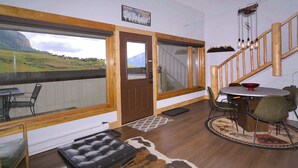 Ein Zimmer mit einem großen Fenster, einer Holztreppe, einem Esstisch mit Stühlen und einem Ledersitzhocker.
