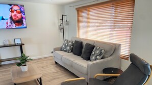 Ein modernes Wohnzimmer mit einem grauen Sofa, zwei Sesseln, einem Couchtisch, einem an der Wand montierten Fernseher und einem Fenster mit Jalousien.
