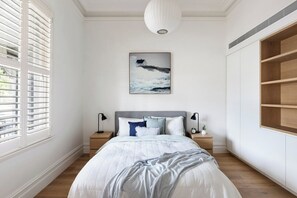 A bedroom with a bed, bedside tables, a nightstand, a wall-mounted painting, and a large window with blinds.