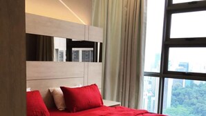 Ein modernes Hotelzimmer mit einem großen Bett, roter Bettwäsche, einem Kopfteil mit integrierter Beleuchtung, Blick auf die Stadt durch große Fenster und einem ordentlich bezogenen Bett mit Handtüchern.
