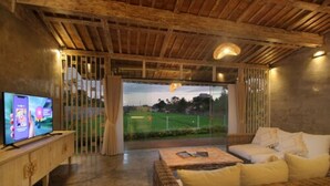 A room with a television on a wooden console, a wicker sofa, and a view of a green field through a large window. - Casa Batu Belig (Keroboken Kelod)