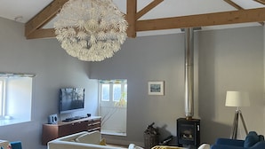A spacious living room with a high ceiling, wooden beams, a large chandelier, a television, a fireplace, and a dining area with a table and chairs. - The Snug - Beautiful Grade 2 listed barn conversion (Mevagissey)