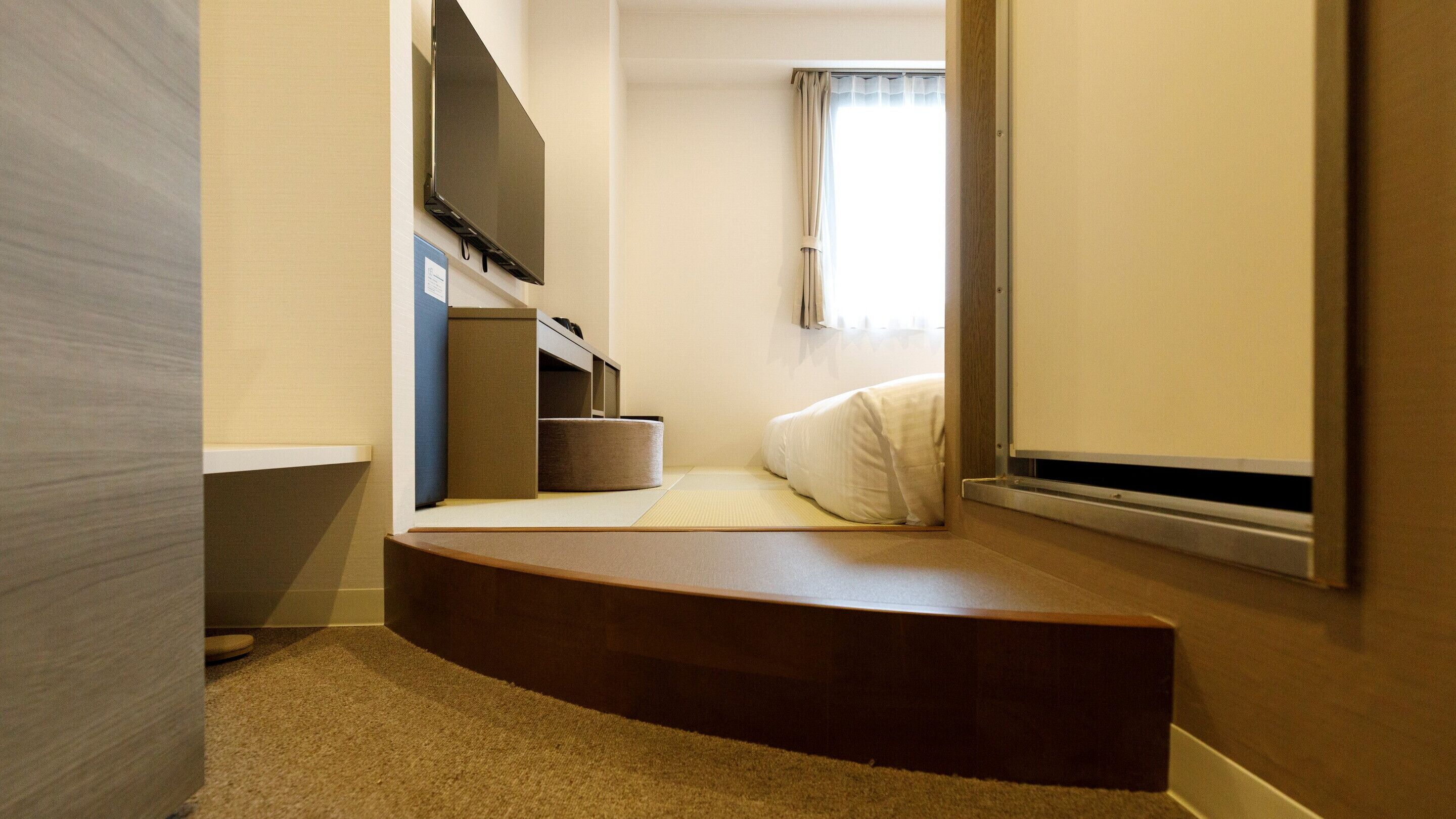 Un dormitorio compacto con televisión integrada, una pequeña zona de estar y una ventana con cortinas.