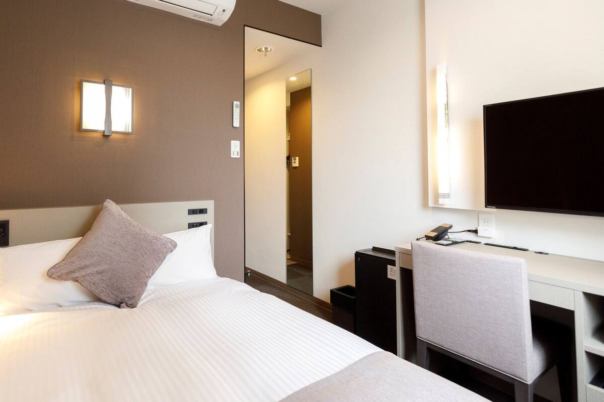 A hotel room with a bed, a desk, a chair, a flat-screen TV, and a mirror.