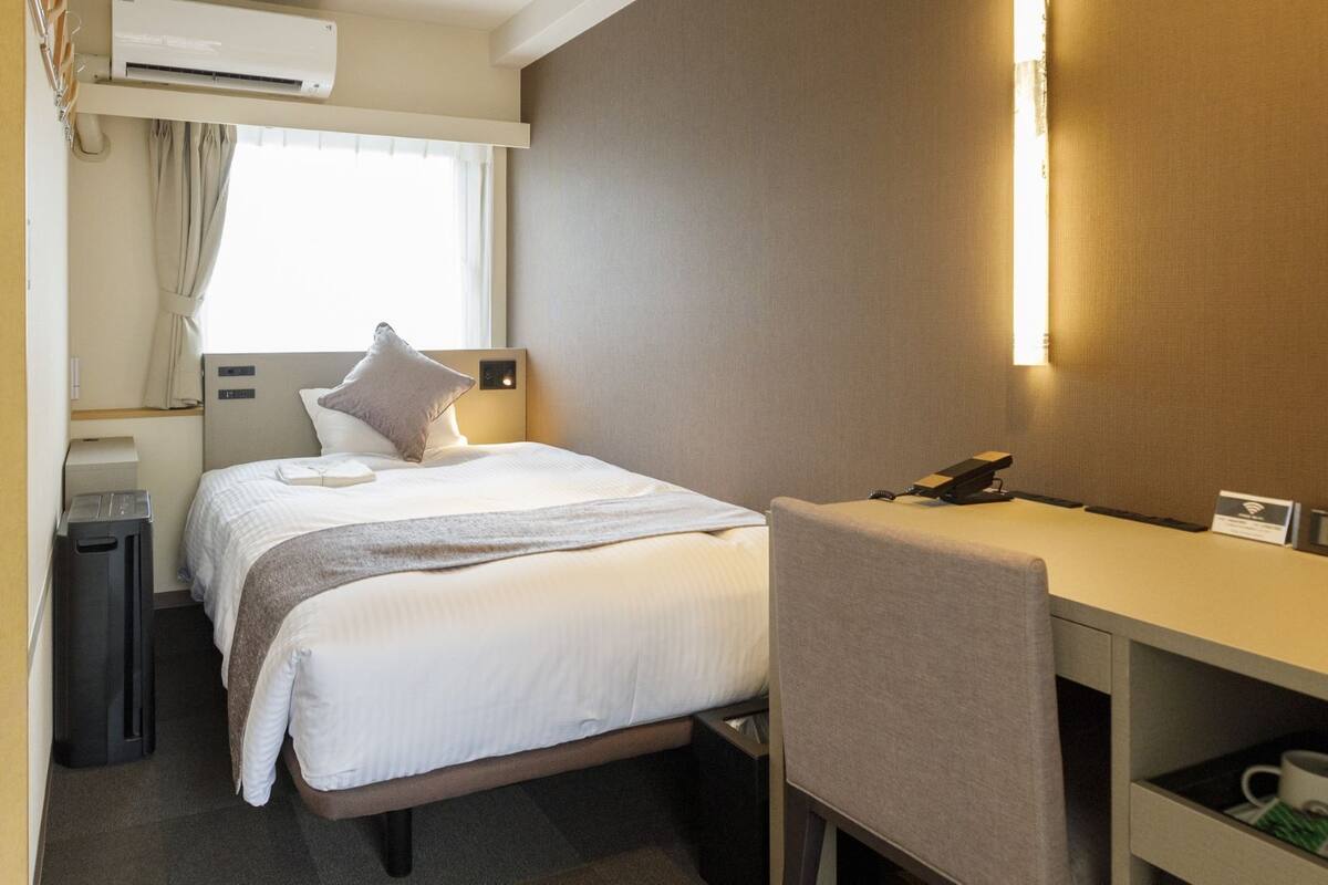 A hotel room with a bed, bedside table, chair, desk, and a small shelf with mugs.