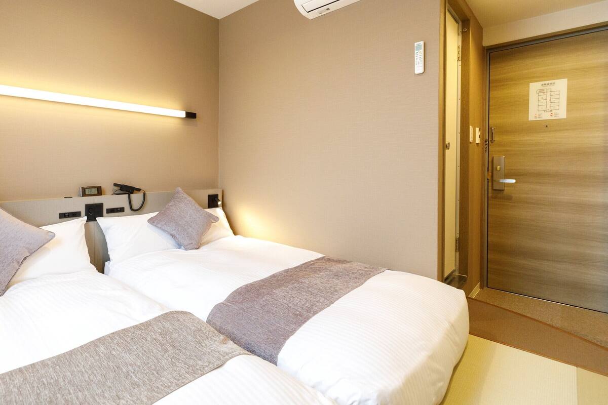 A hotel room with two beds, a bedside table with a phone, a wall-mounted light, and a door with a sign.