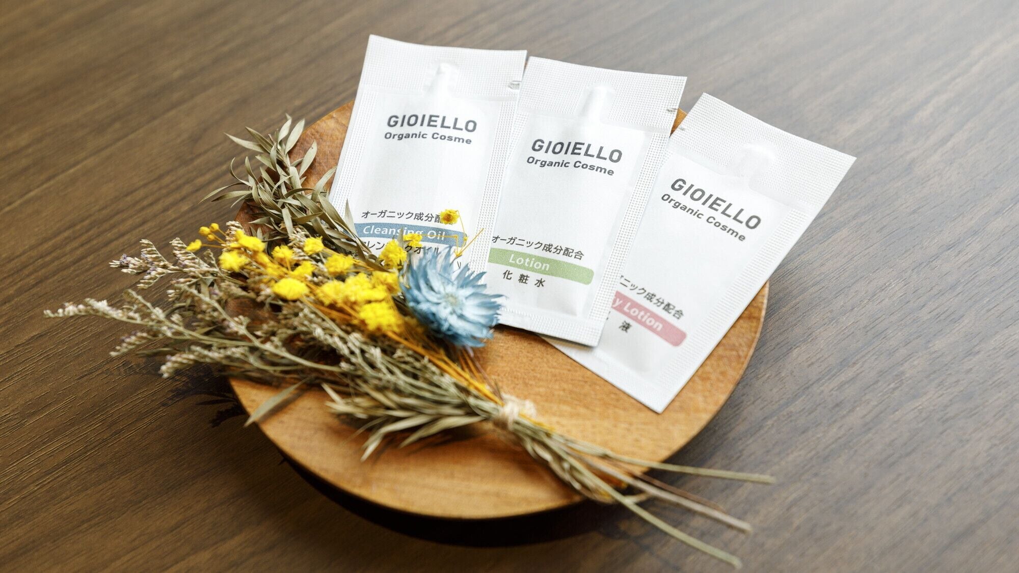Três produtos de cosmética orgânica GIOIELLO sobre uma bandeja de madeira com flores secas.