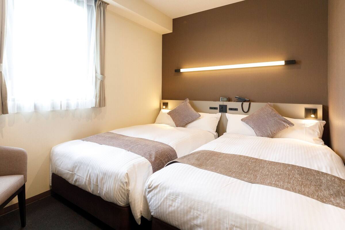 A hotel room with two beds, a chair, and a bedside table.