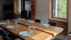 Un comedor con una larga mesa de madera dispuesta para seis personas, flanqueada por sillas negras. A la izquierda, un mueble negro con un televisor, y a la derecha, una ventana con vista a la vegetación.