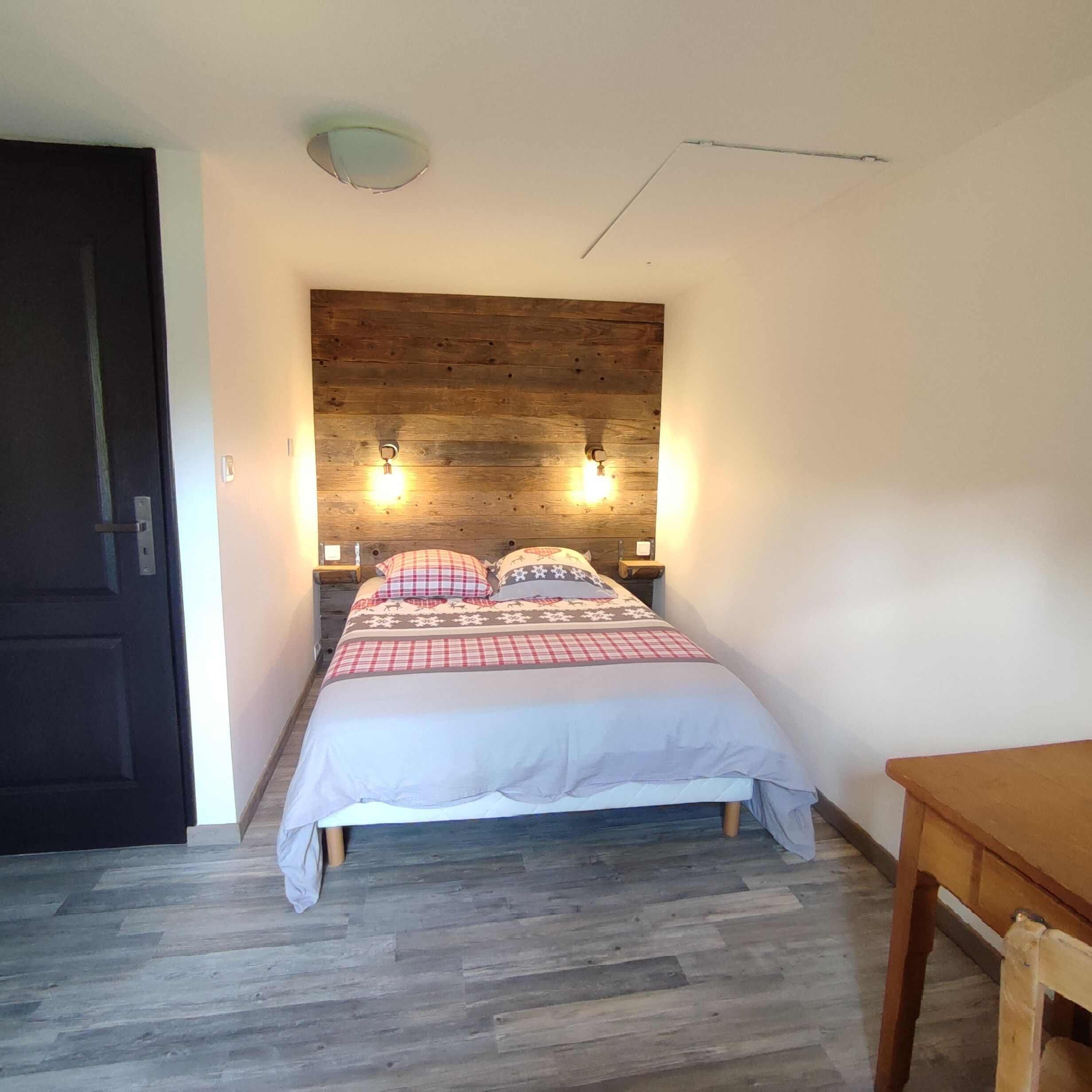 Un dormitorio con cabecera de madera, cama con colcha a cuadros, dos lámparas de noche y una mesita de madera.