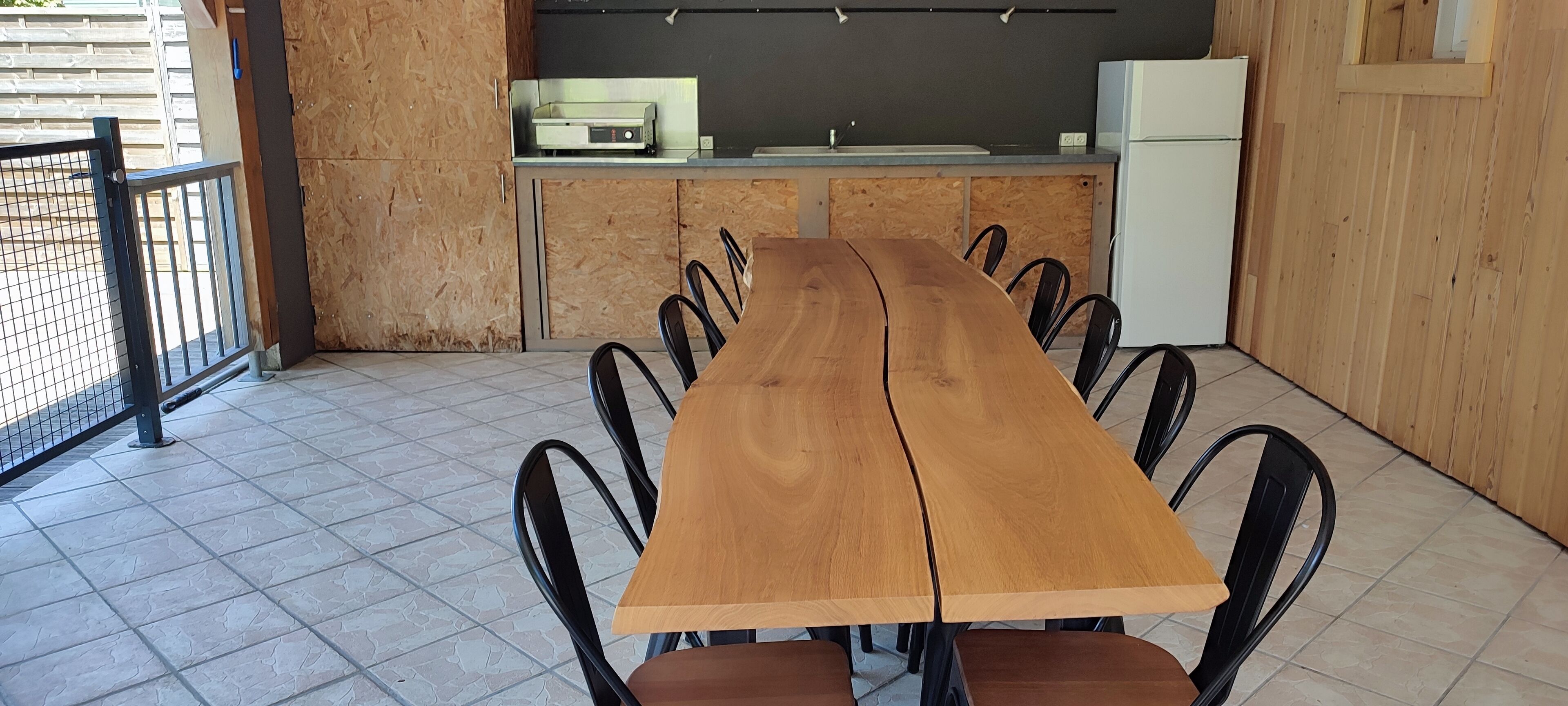 A long wooden table with black chairs in a room with a tiled floor, a kitchen area with a refrigerator and a microwave, and a wooden wall.
