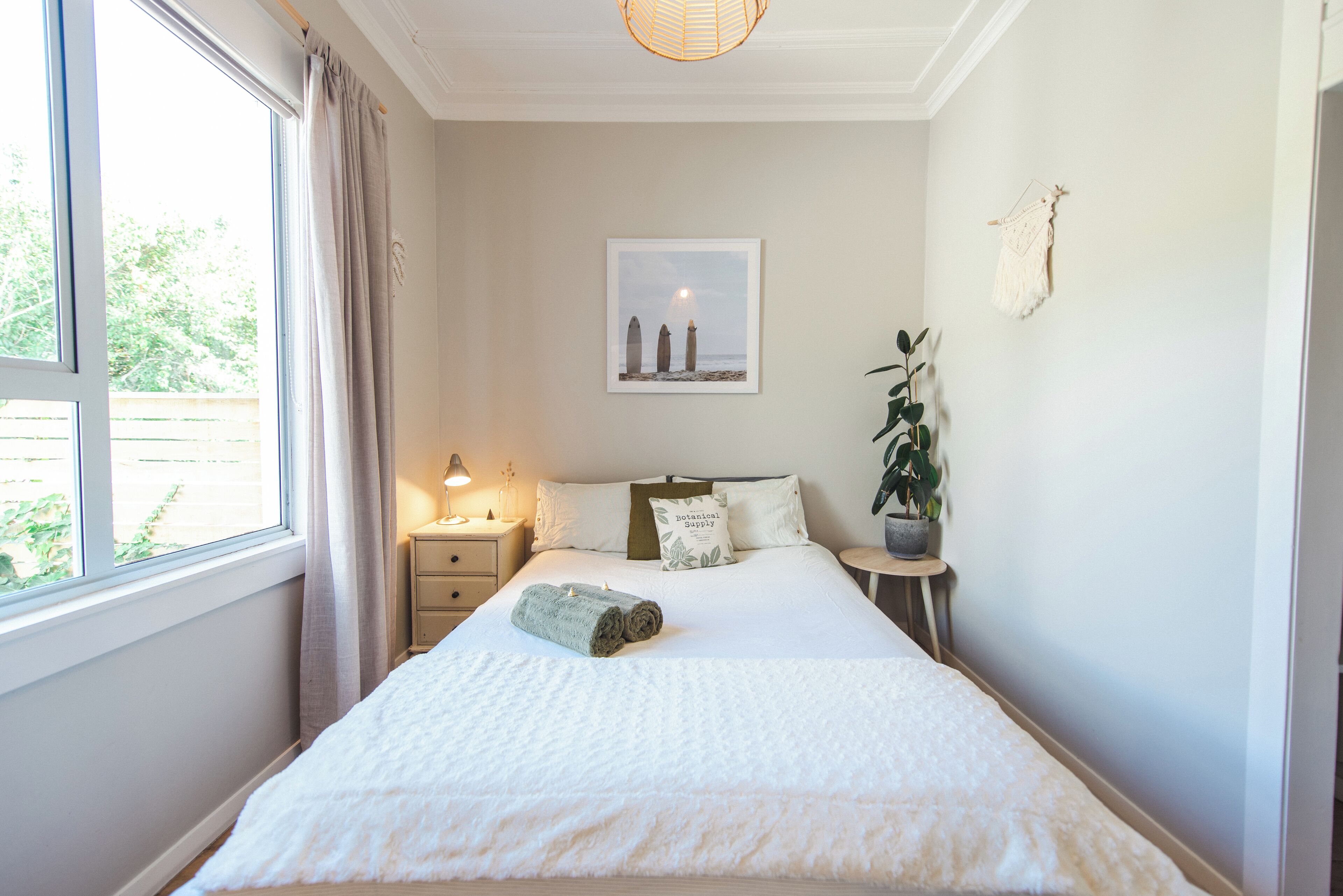 A neatly made bed with a rolled towel, a bedside table with a lamp, a framed picture on the wall, and a potted plant.